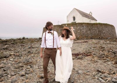 Bride and groom walking hand in hand away from St. Cwyfan's, The Church in the Sea in Aberffraw, by Anglesey Photographer Gill Jones Photography