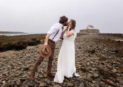 Groom leaning forward to catch a kiss from his new wife, they're standing on a causeway with St. Cwyfan's, The Church in the Sea in Aberffraw in the background by Anglesey Photographer, Gill Jones Photography